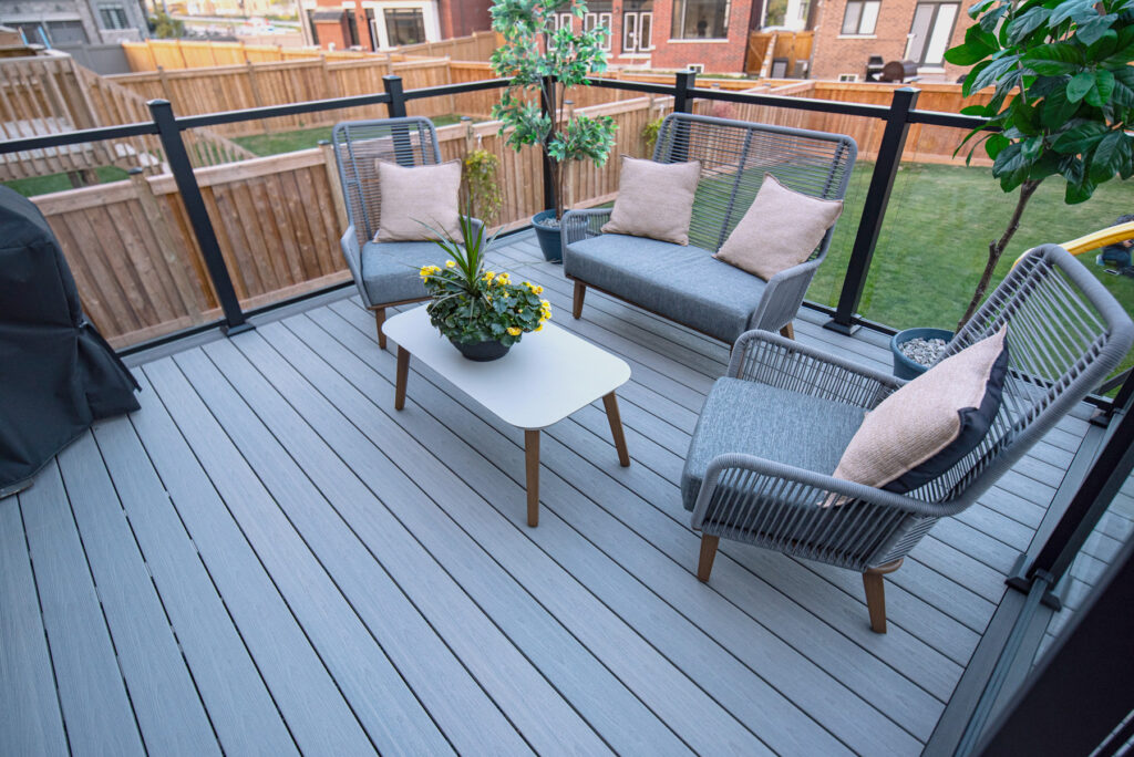 A modern Fiberon Chai deck features gray flooring and a seating area with four gray chairs, each adorned with a beige cushion. A white rectangular table holds a potted plant with yellow flowers. The deck, encompassed by Fortress Railings4 and a wooden fence, offers contemporary charm.