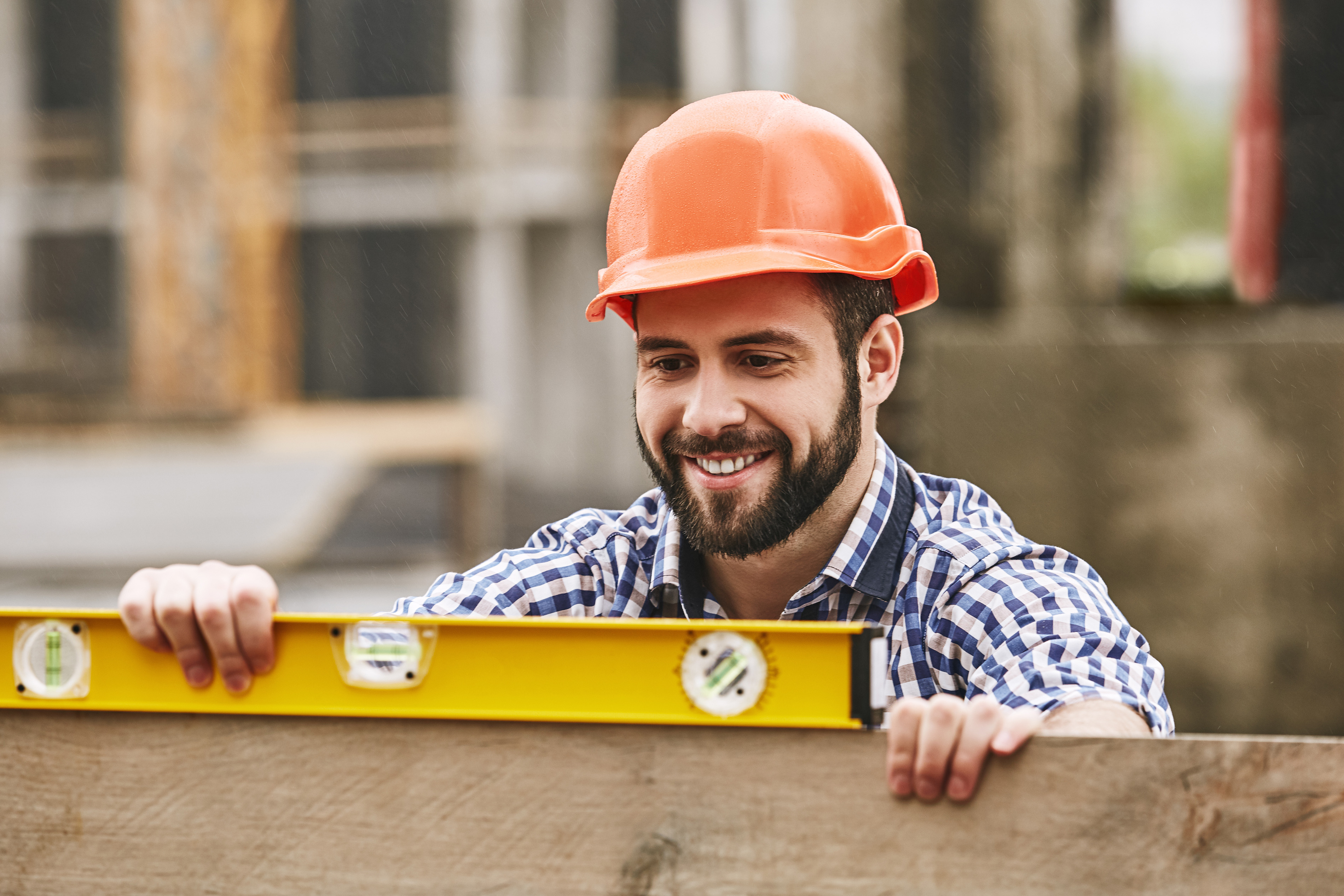 A man in a checkered shirt and orange hard hat uses a yellow level tool to ensure the perfect alignment of a wooden plank at the construction site. As a Custom Composite Deck Builder in Toronto, he is smiling and fully engaged in his meticulous work.