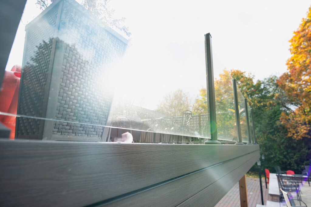 Sunlight beams through a patio lined with wicker planters and a glass railing, expertly installed by Railing Installer Toronto. The bright glow casts over autumn trees with colorful leaves, framed against the clear sky in the background.