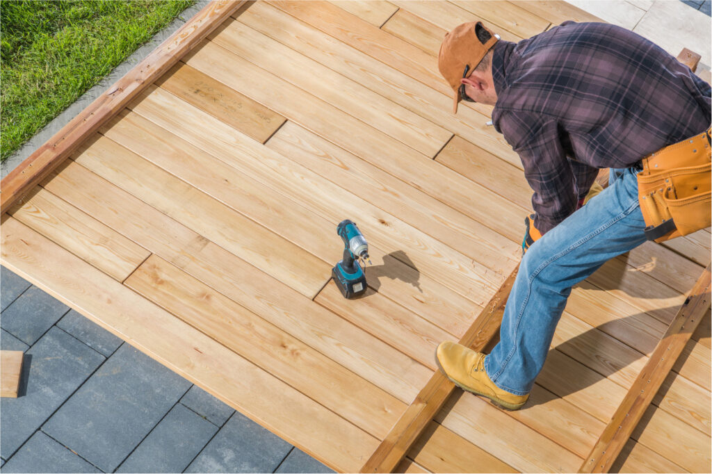 A person wearing a brown cap and tool belt is kneeling while constructing a wooden deck. They are surrounded by wooden planks and a power drill. The deck is next to a grassy area and a paved walkway.
