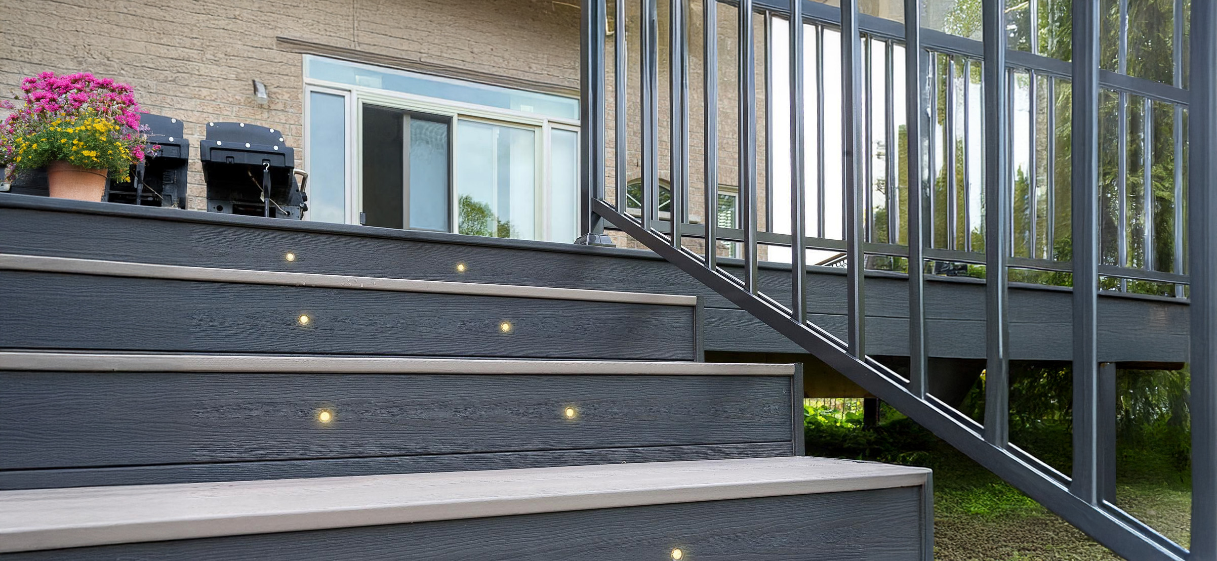Deck_Railings-and-Lights-installer Outdoor stairs, adorned with built-in lights, lead up to a deck, where sleek metal railings flank the steps. A potted plant with pink flowers sits near a glass sliding door that opens to the house, while a barbecue grill awaits on the well-lit deck.