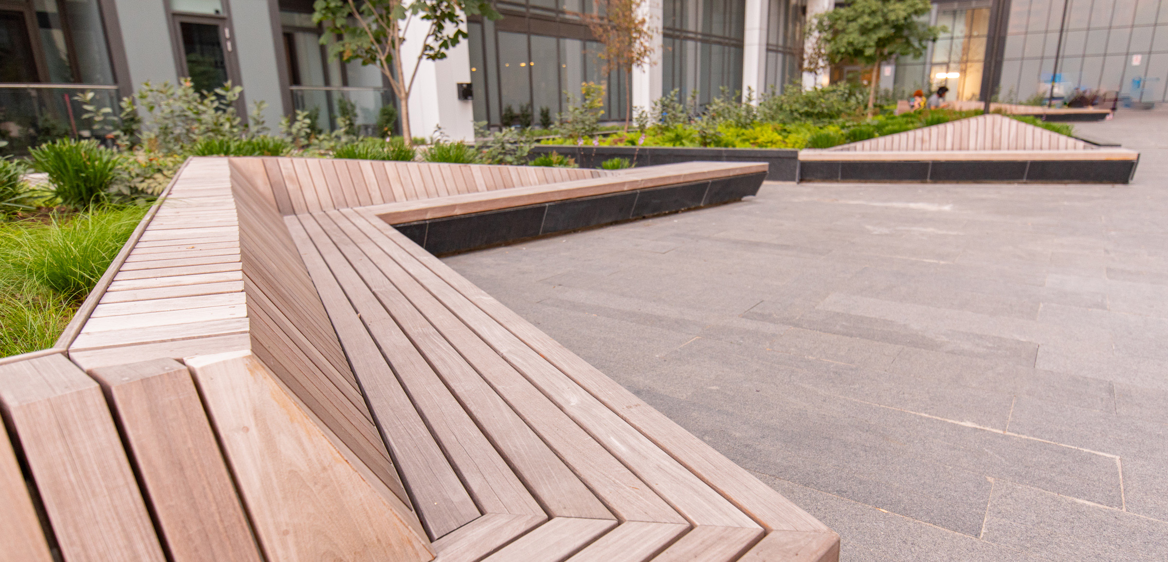 IPE_benches_condo_yard Wooden benches arranged in angular, geometric shapes are set against a contemporary building backdrop. The area features greenery and trees integrated into the design, with large windows and paving stones creating a modern outdoor space.