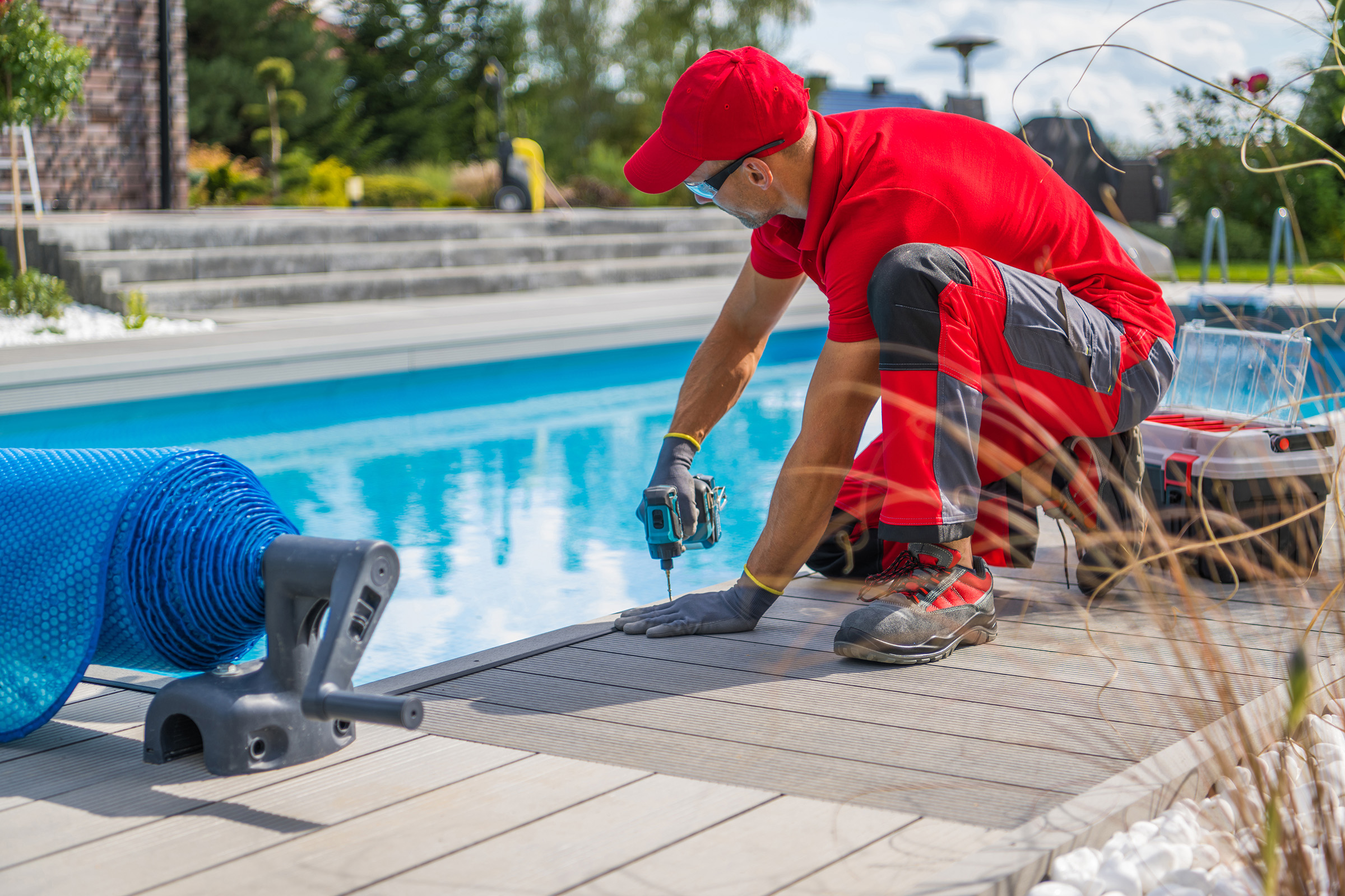 A pool deck contractor in Toronto, dressed in red clothing and gloves, kneels by the pool, expertly using a power drill on the deck. Nearby, a roll of blue pool cover and a toolbox lie under the sunny sky amidst steps and greenery.