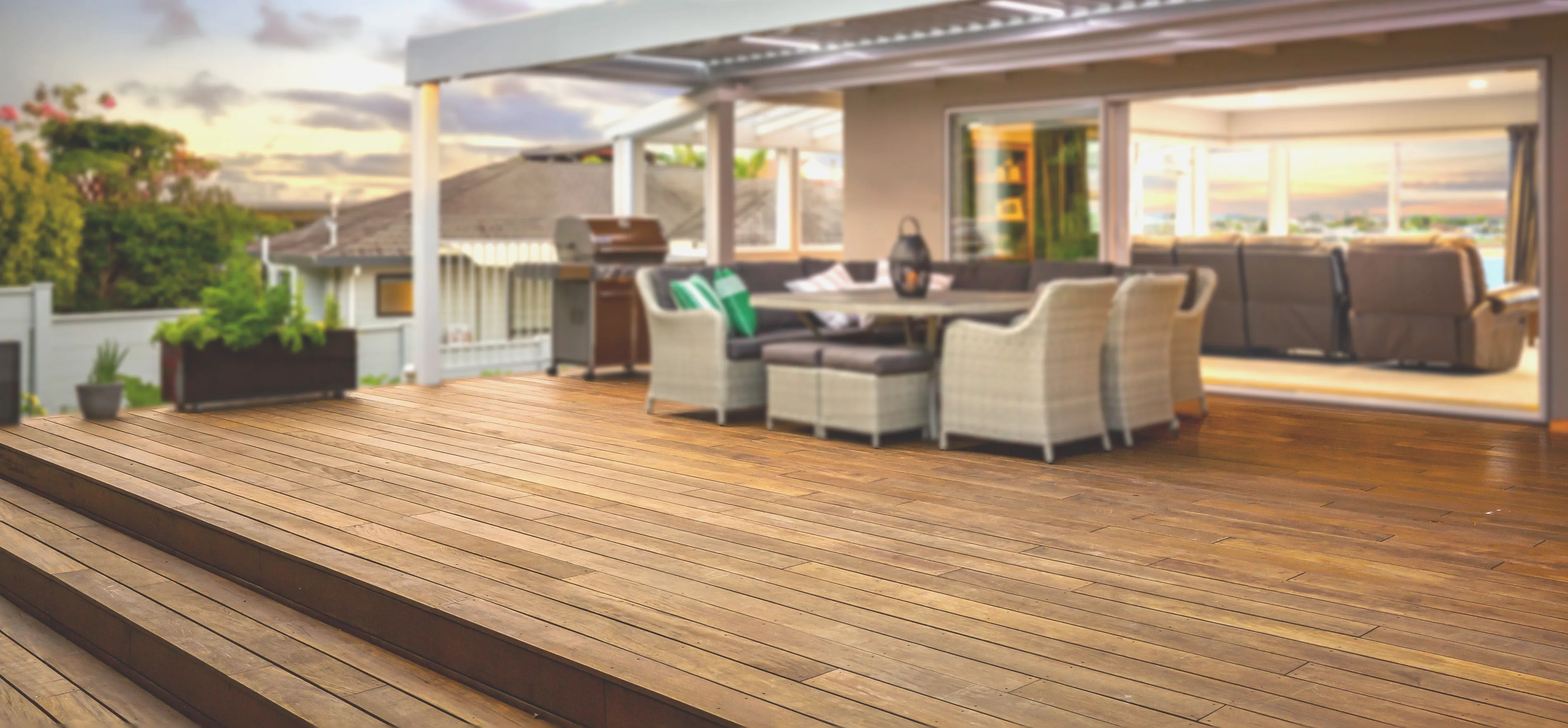 Rooftop-Decking-Balcony-Toronto Spacious wooden deck crafted by a renowned Deck Builder Toronto, featuring an outdoor dining set under a pergola, adjacent to a contemporary home. Open sliding doors reveal a cozy interior with sofas. Lush greenery surrounds the area as the sky is partly cloudy at sunset.