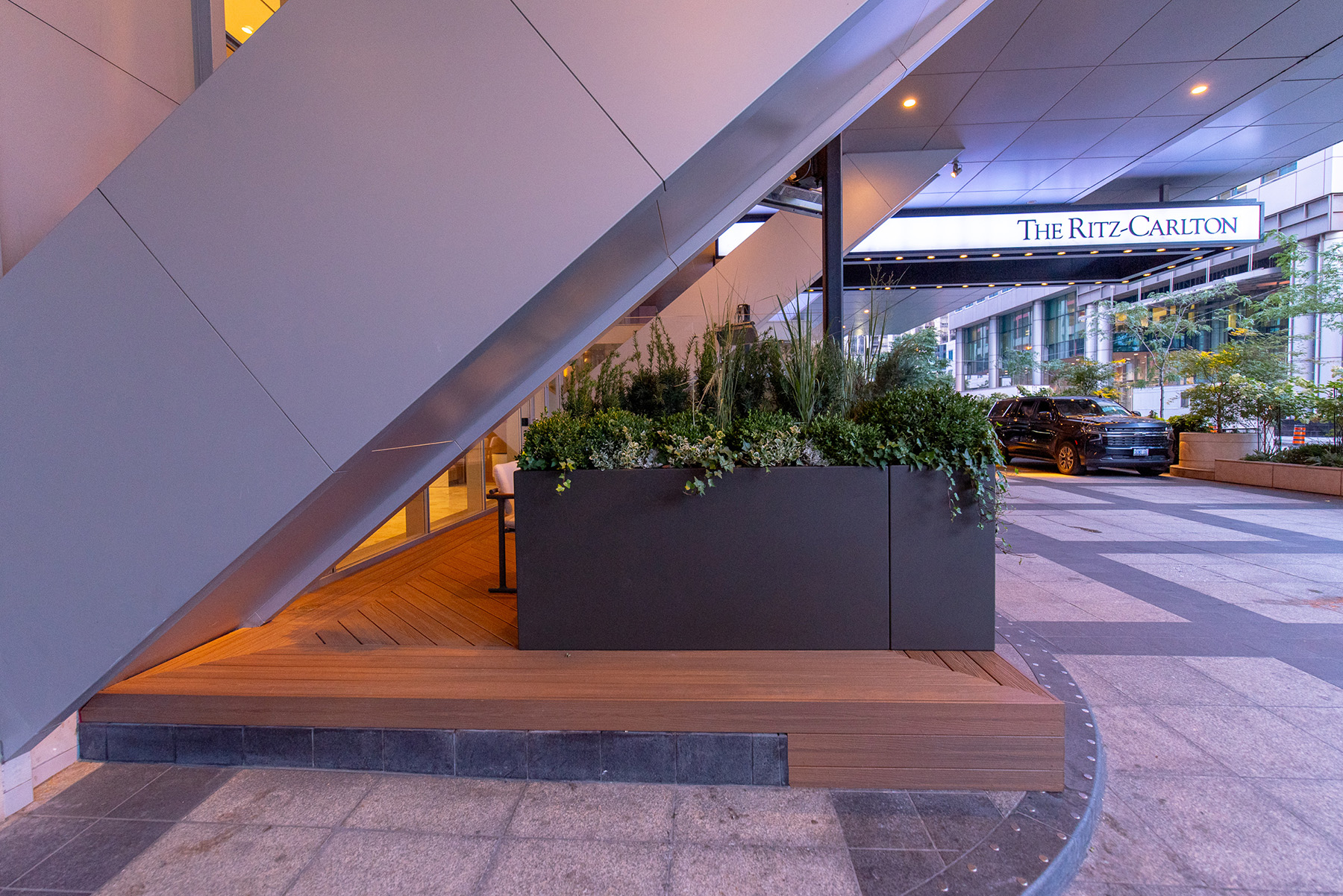 A modern hotel entrance features a wooden deck and large planter with greenery. The Ritz-Carlton sign is visible in the background, and a black car is parked nearby. The architecture includes sleek metal panels and glass elements.