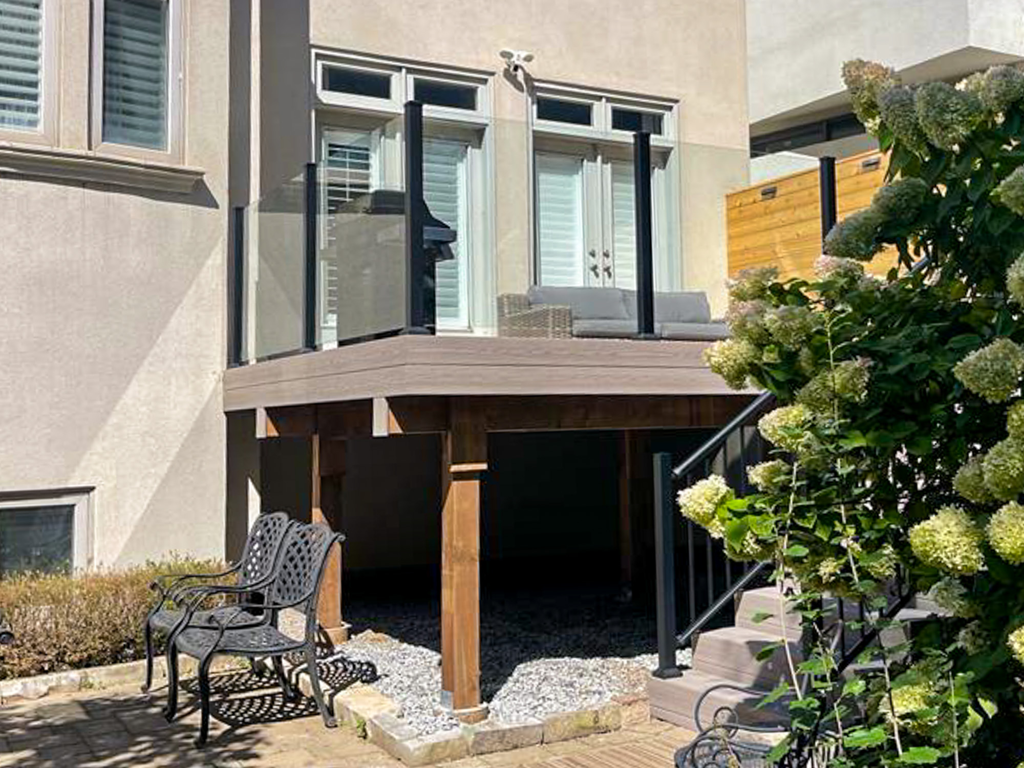 A modern backyard patio with glass railings, a small outdoor sofa, and wooden support beams. A metal chair and flowers are in the foreground, and stairs lead up to the patio. Sunlight casts shadows across the area.