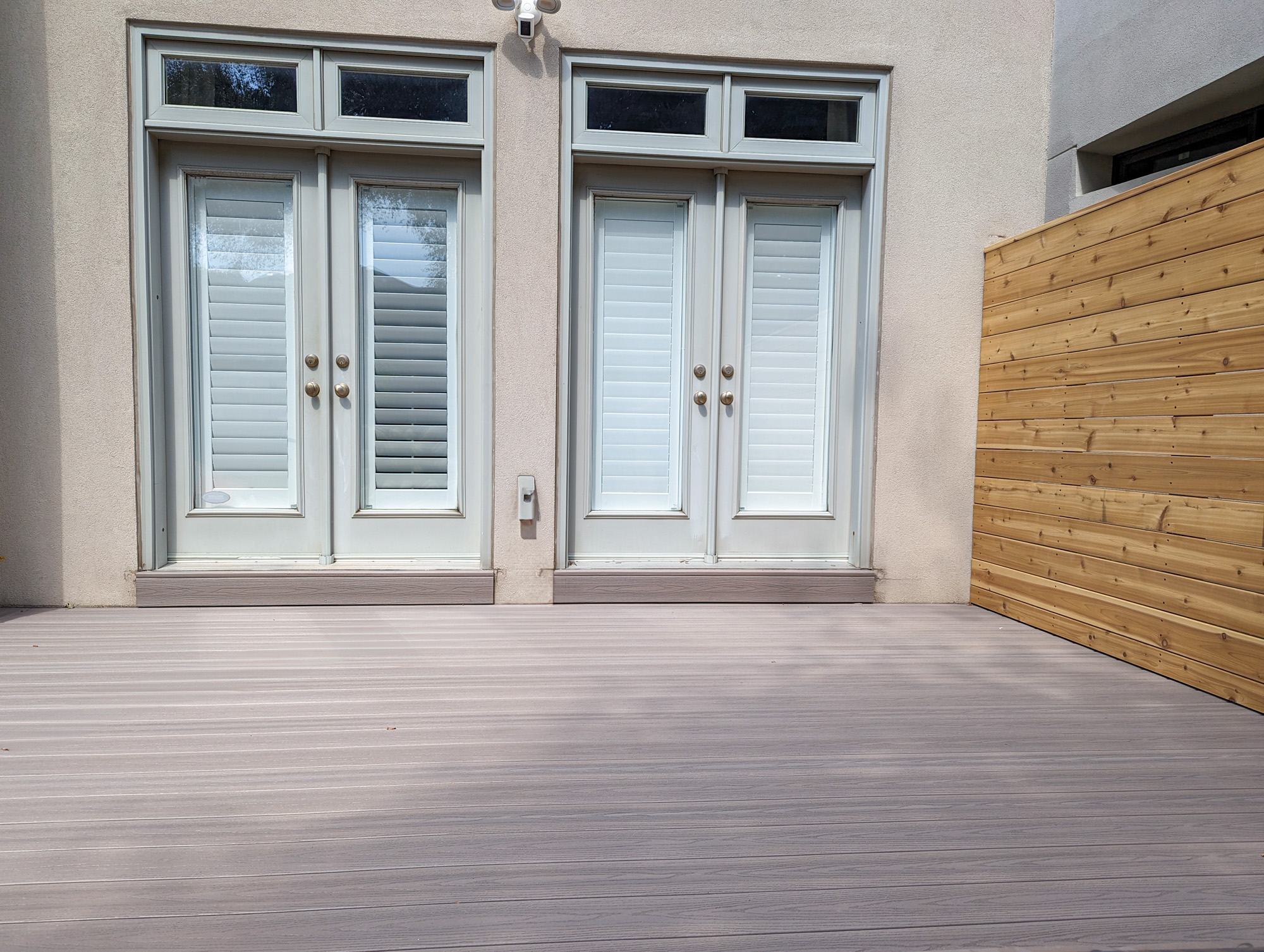 Two white double doors with closed shutters lead to a light gray patio deck. A wooden privacy fence is on the right side, and the wall and surroundings are painted in neutral colors.