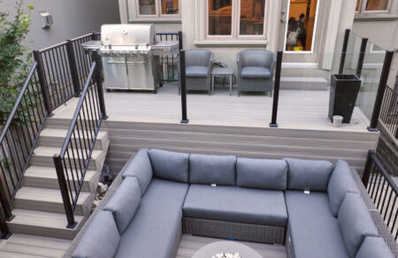 A modern outdoor patio with Fiberon Composite Decking features a sunken seating area where gray cushioned wicker sofas form an L-shape around a round coffee table, surrounded by stairs and sleek black railings.