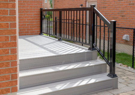 A small set of light gray steps with a matching platform, bordered by a black metal railing, leads up to a brick house with a fenced yard visible in the background.
