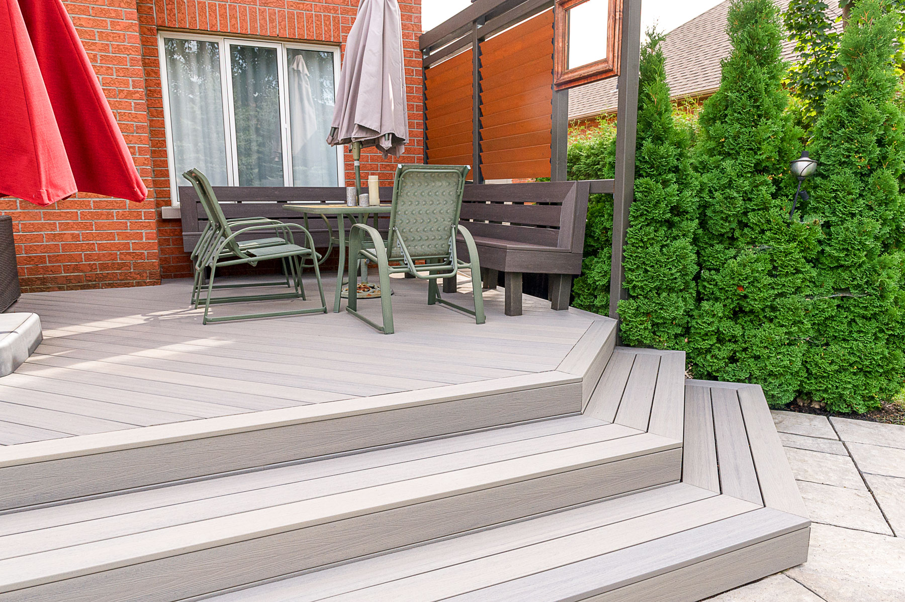 A modern backyard patio with gray composite decking, green chairs, a bench, a table, large red and gray umbrellas, and privacy fencing, surrounded by green hedges and a brick house.