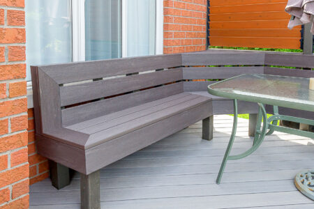 A modern gray wooden corner bench is positioned next to a glass-top patio table on a deck with light gray flooring, adjacent to a brick wall and a window.