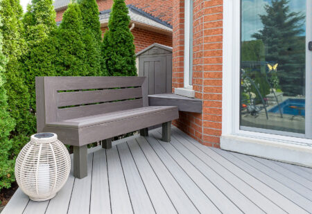 Gray wooden bench and white lantern on a light gray deck next to a red brick house, with tall green shrubs and a glass door reflecting an outdoor pool area.