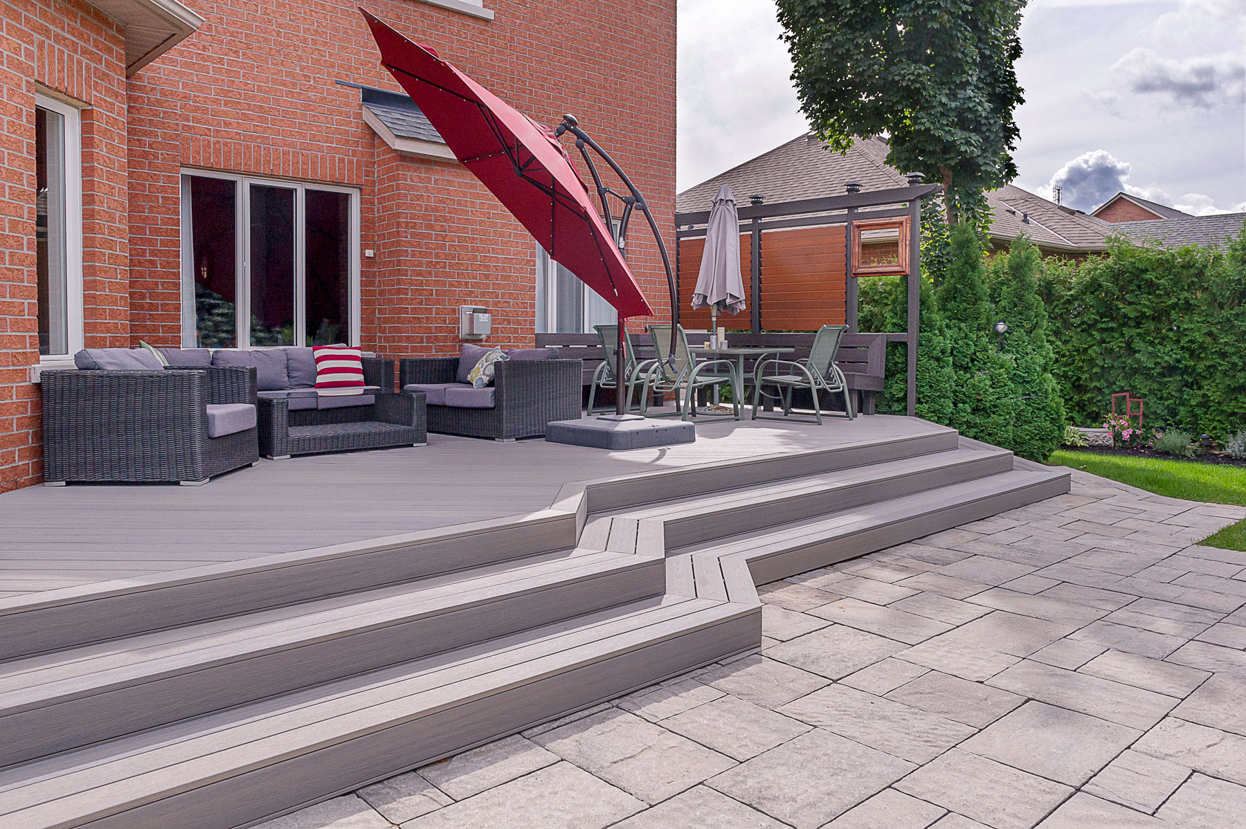 A backyard patio featuring gray composite decking, outdoor sofa set, red umbrella, dining table with umbrella, and stone paving. A brick house and lush greenery create a charming backdrop.