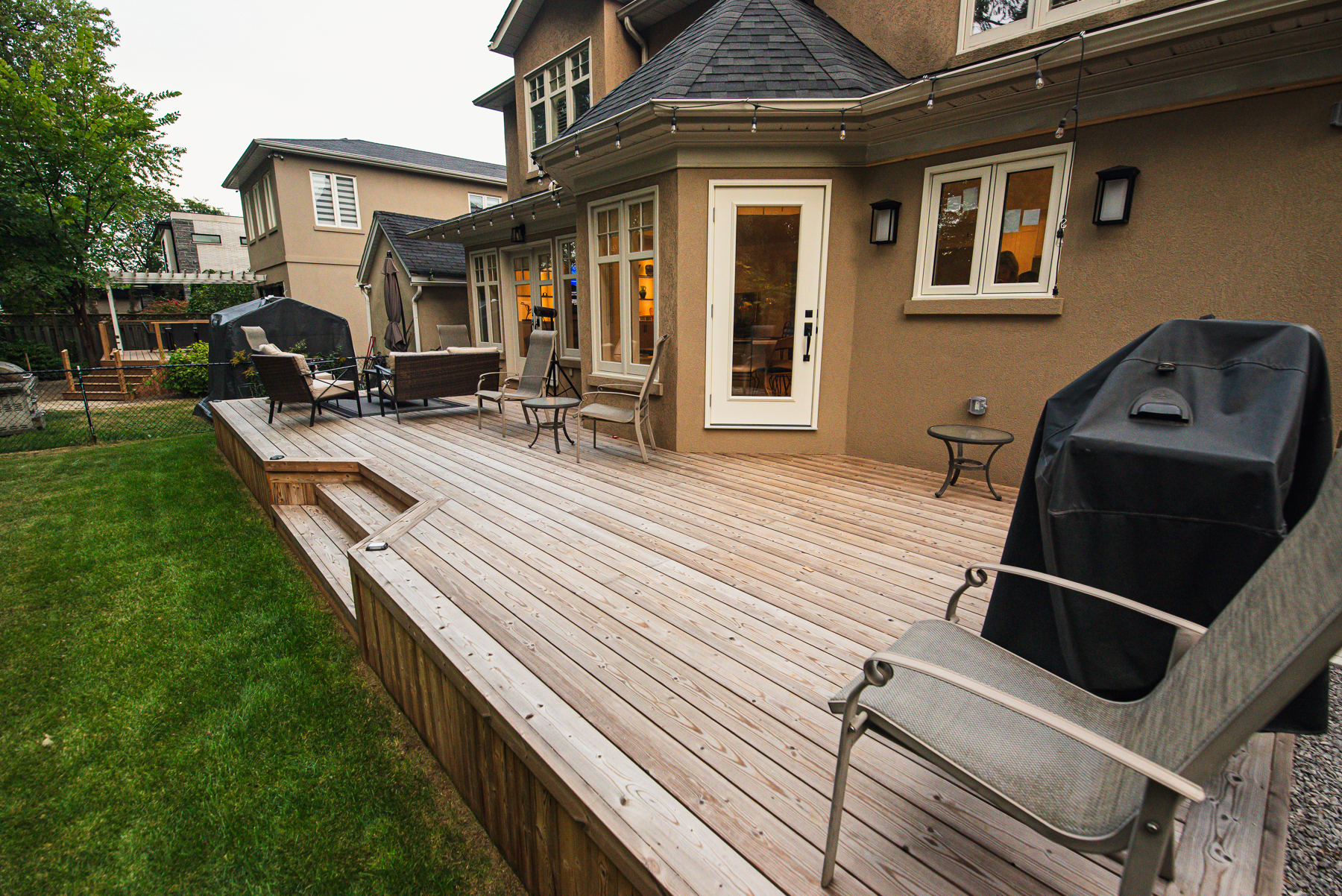 A spacious Lunawood Thermowood Deck with outdoor furniture and a covered grill, attached to a beige house with large windows and a glass door, overlooks a lush green lawn.