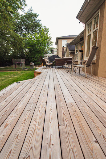 A spacious wooden backyard deck with Lunawood Decking features patio chairs and tables, attached to a house. The deck overlooks a grassy yard with trees and fencing, creating an inviting outdoor seating area.