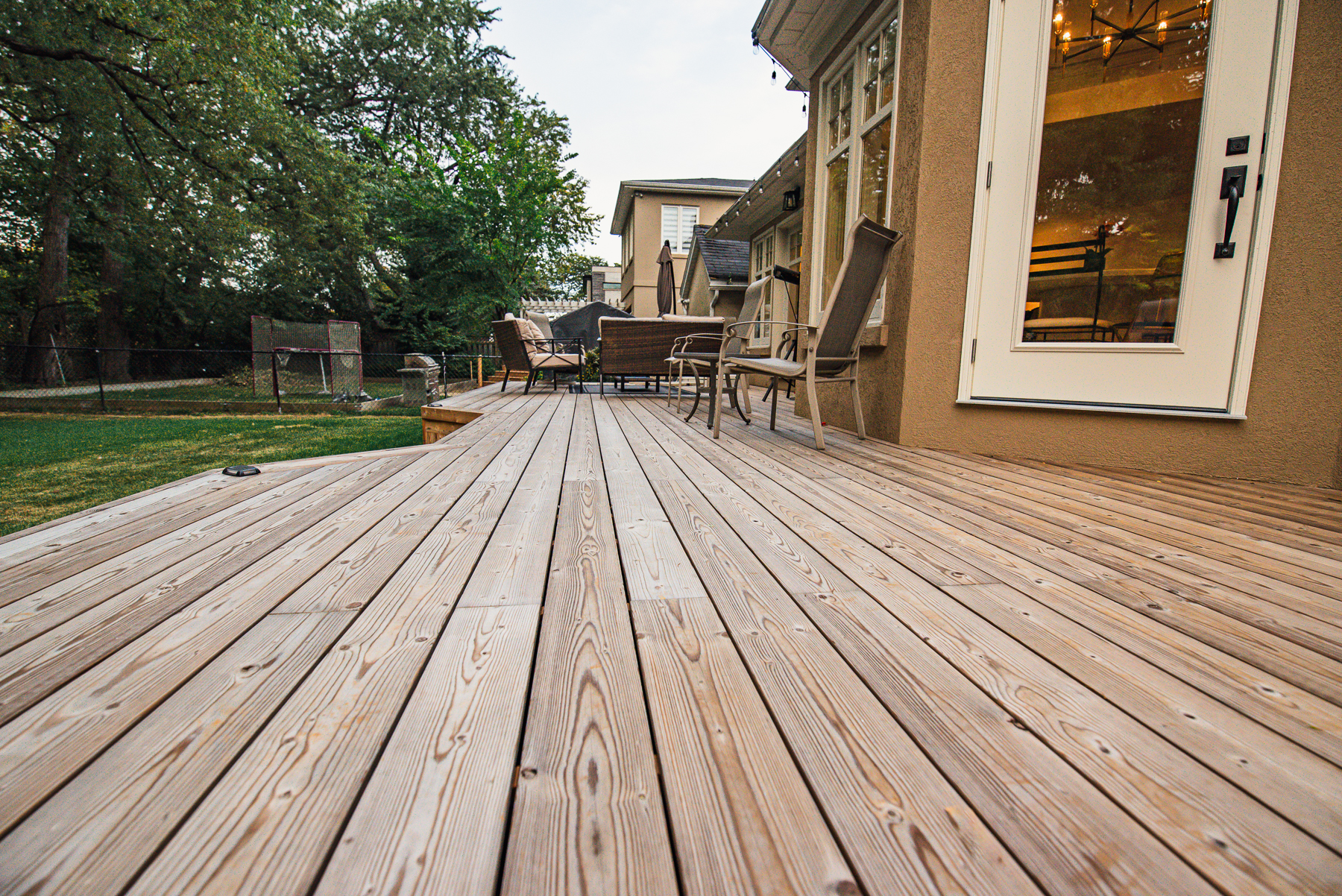 A spacious Lunawood Decking attached to a house, featuring several chairs and a table for outdoor seating. The deck overlooks a grassy backyard dotted with trees and fenced for privacy.