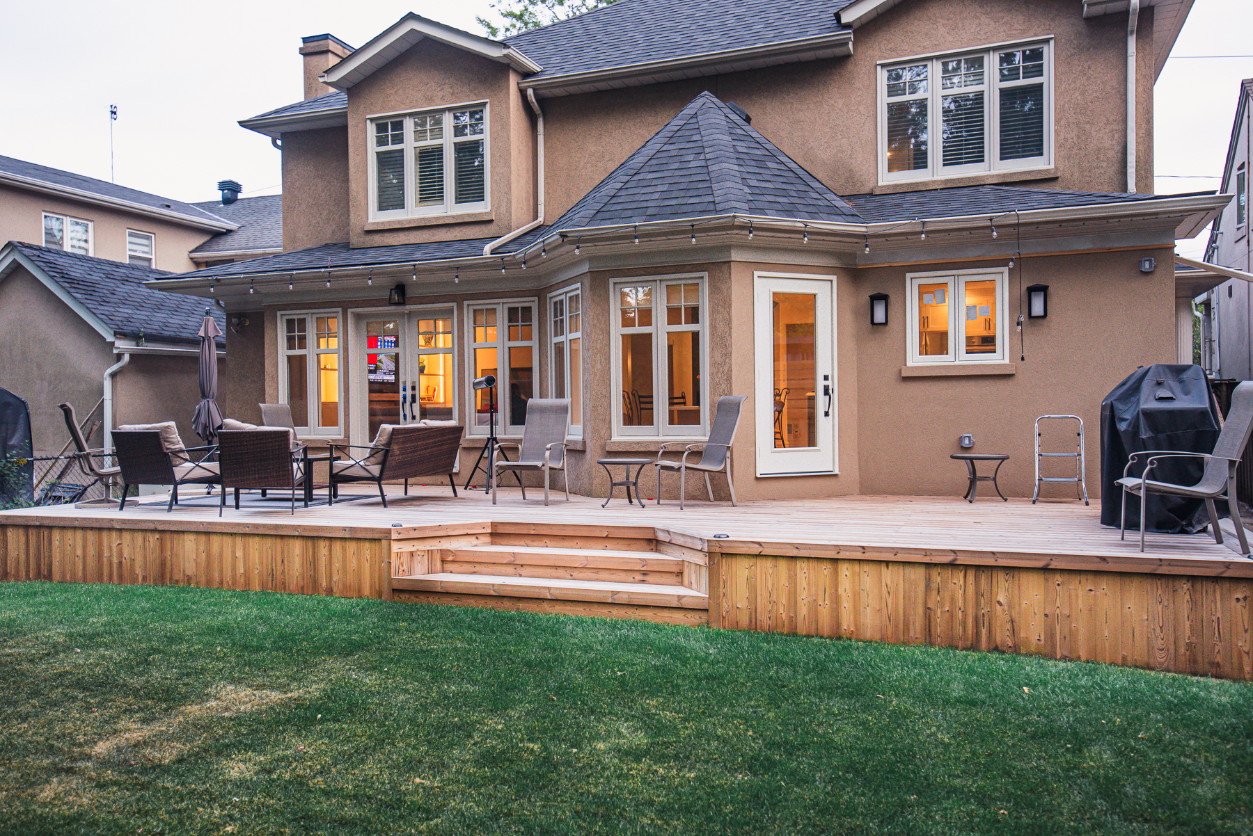 A spacious Lunawood backyard deck with outdoor chairs, tables, and a grill, attached to a two-story tan house with many windows and a well-kept lawn.
