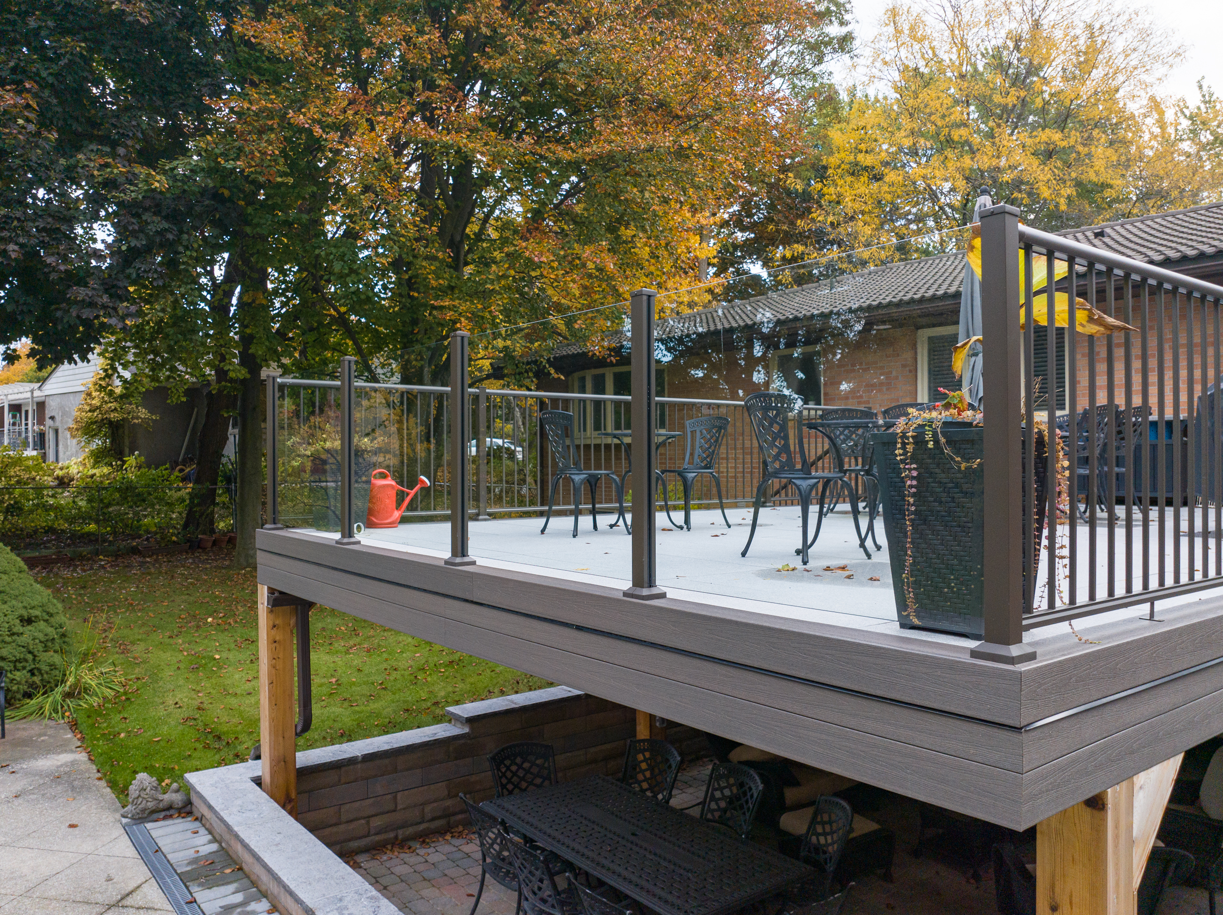 A raised backyard deck with Century Scenic (glass front) railings holds black patio furniture and a red watering can. Below, there’s a shaded patio area with a table and chairs, all surrounded by trees with autumn foliage.