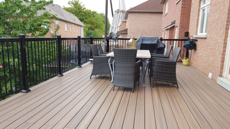A spacious backyard deck with brown PVC decking, black railing, a dining table with six wicker chairs, a barbecue grill, potted plants, and views of neighboring houses and trees in the background.