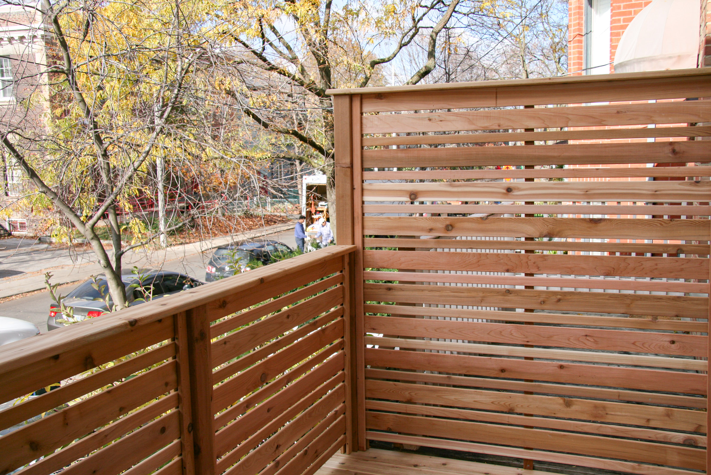 A small wooden balcony with horizontal slats overlooks a street with parked cars and trees losing their leaves.