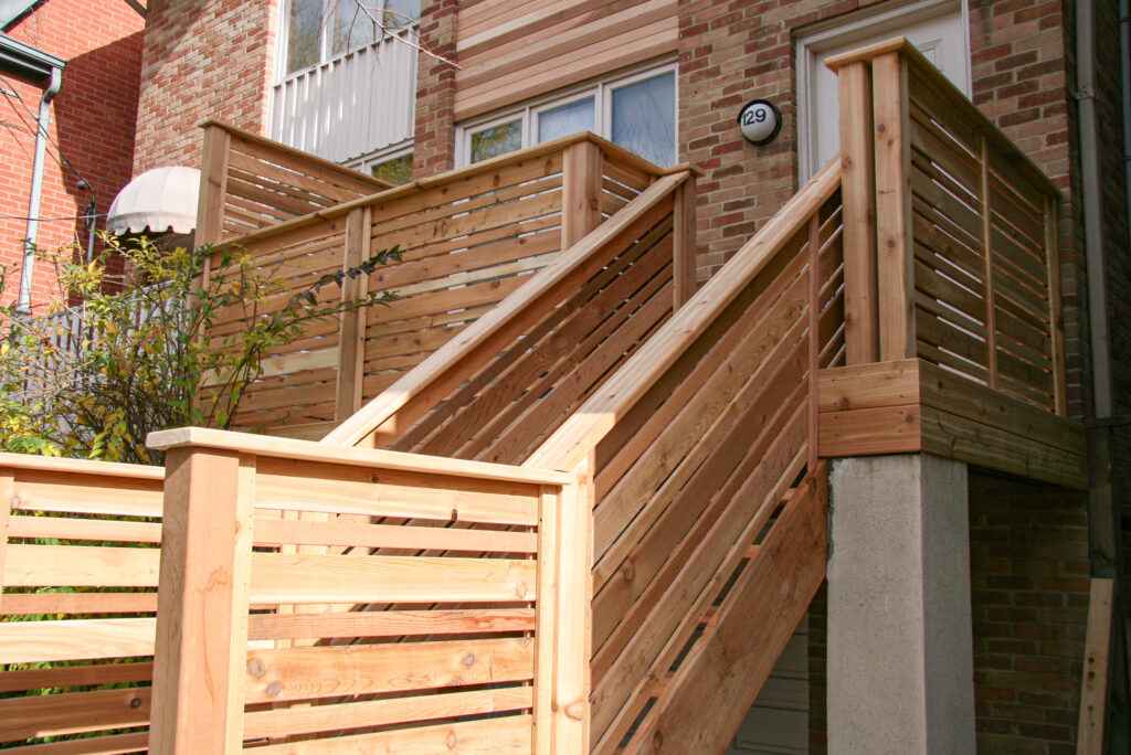 A townhouse with a brick exterior, cedar balcony, and staircase leading to a white front door on the second level.