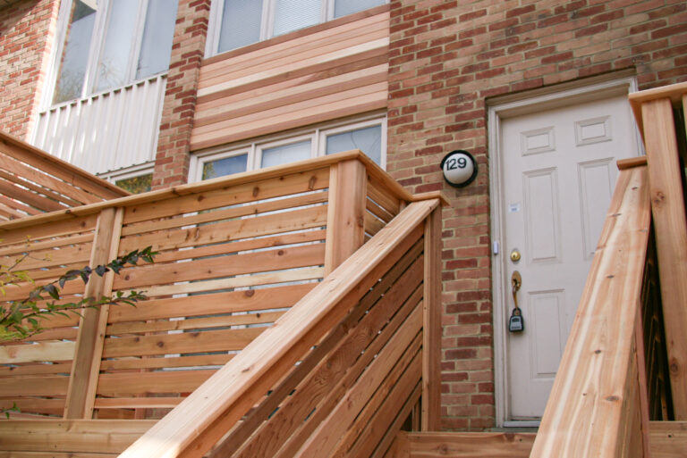 A beige front door with a lockbox, surrounded by a new wooden railing and brick exterior, under a sign reading 129.
