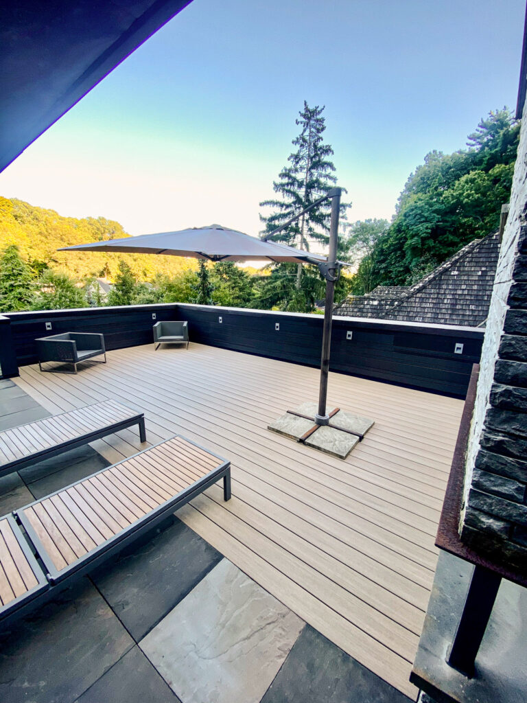Spacious outdoor patio with lounge chairs, a large umbrella, and surrounded by trees under a clear sky.