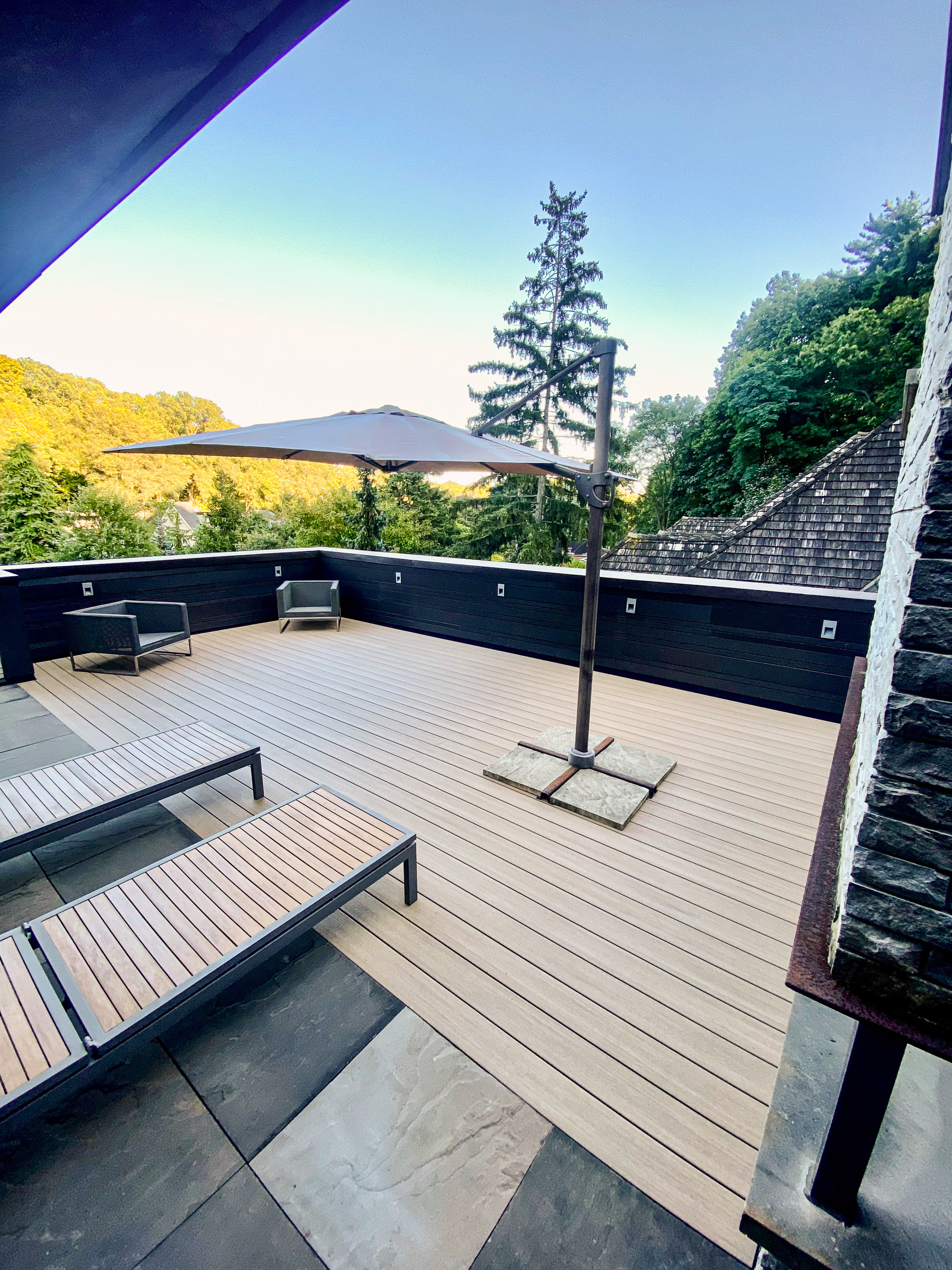Spacious outdoor patio with lounge chairs, a large umbrella, and surrounded by trees under a clear sky.