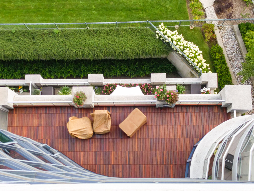 Aerial view of an IPE wooden balcony with two covered chairs, a table, and potted plants overlooking a green lawn.
