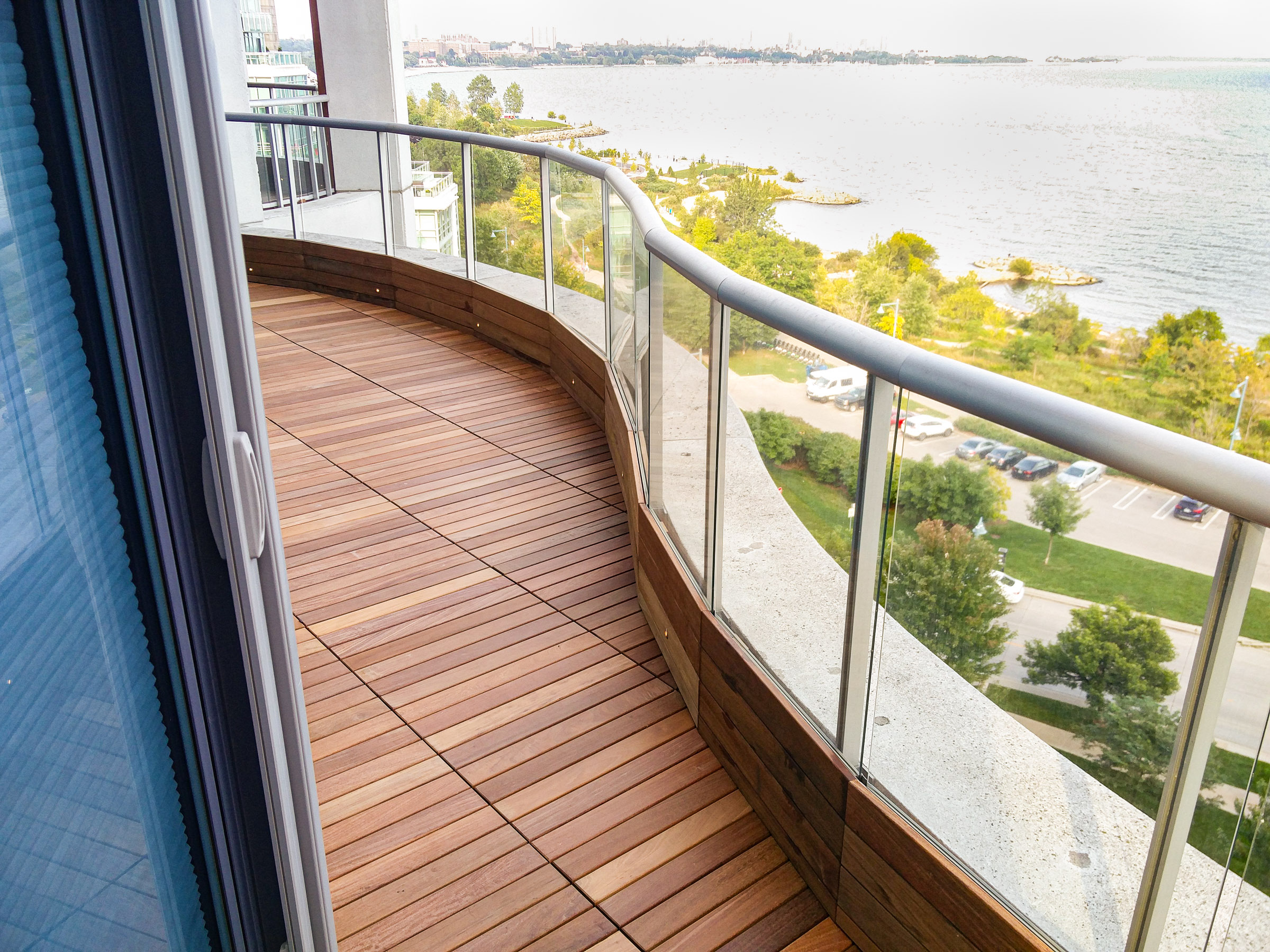 Curved balcony with IPE wooden flooring and glass railing overlooks a park, parking lot, and body of water.