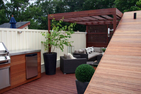 Modern backyard patio with a wood deck, outdoor kitchen, potted plants, and a shaded seating area under a pergola.
