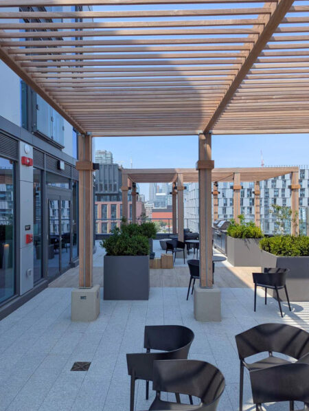 Rooftop patio with wooden pergola, black chairs, tables, and planters, overlooking a cityscape in the background.