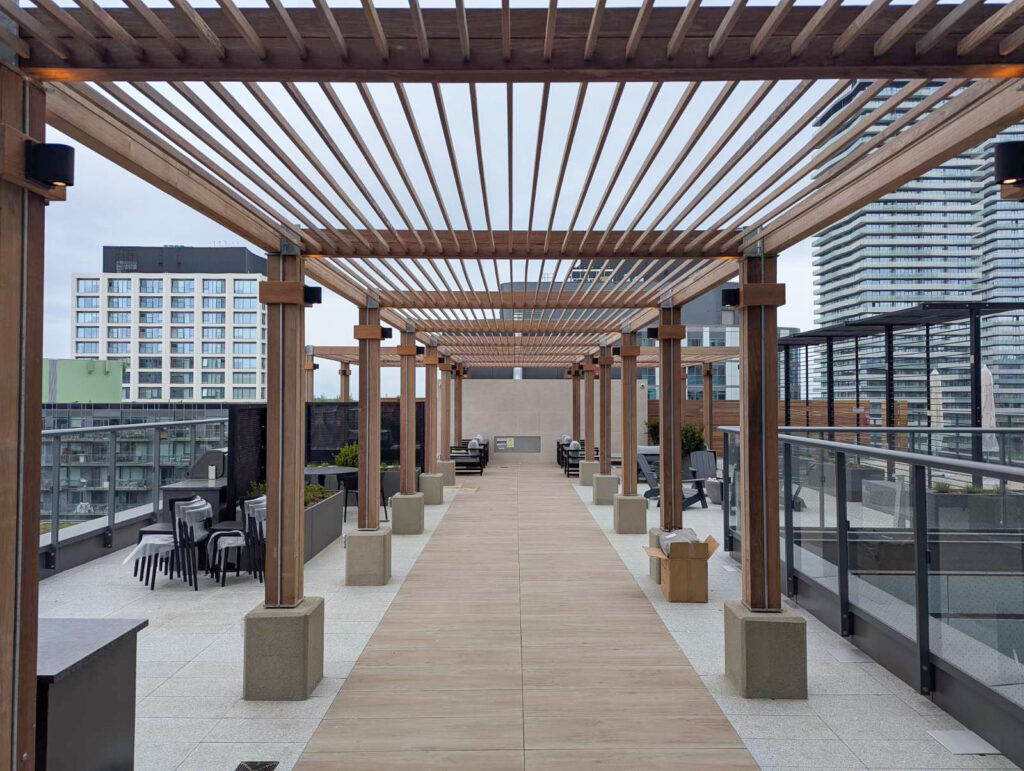 A rooftop patio with wooden pergolas, seating areas, and city buildings visible in the background under a cloudy sky.