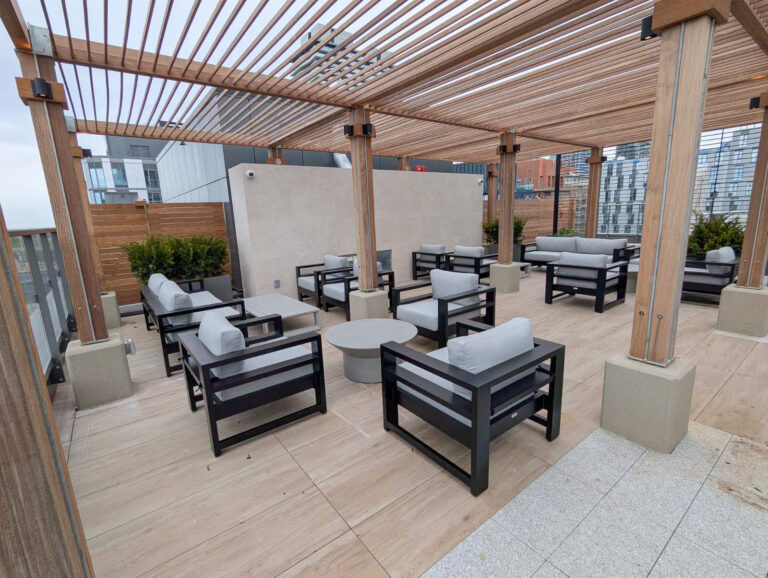 Rooftop patio with wooden pergola, gray cushioned chairs, sofas, and round tables, overlooking modern buildings.