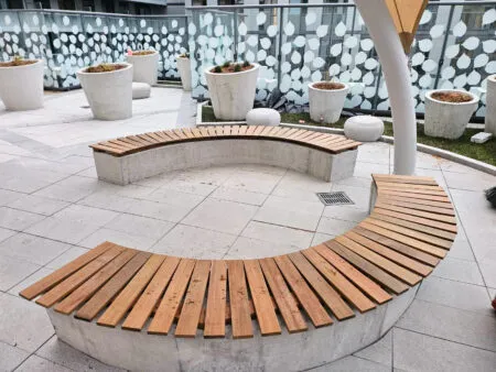 Curved wooden benches with concrete bases in a modern outdoor area with potted plants and a patterned glass railing.