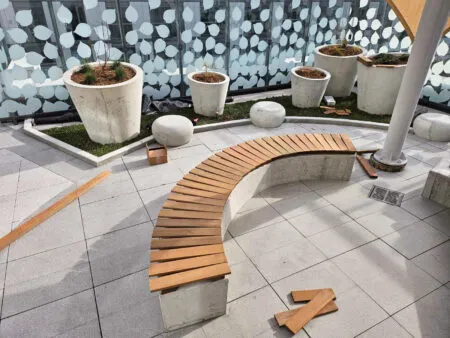 A curved Ipe wooden bench under construction on a tiled patio with large planters and scattered wooden planks.