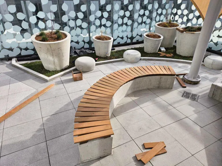 A curved Ipe wooden bench under construction on a tiled patio with large planters and scattered wooden planks.
