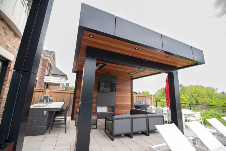 Modern outdoor patio with a wooden ceiling, lounge seating, TV, and built-in grill area next to a fenced yard.