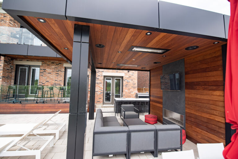 Covered outdoor patio with wood ceiling and walls, lounge seating, TV, fireplace, and a bar area, next to a brick house.