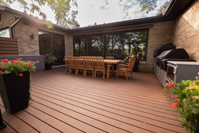 Spacious backyard deck with a long wooden dining table, chairs, potted flowers, and a grill next to a brick house.