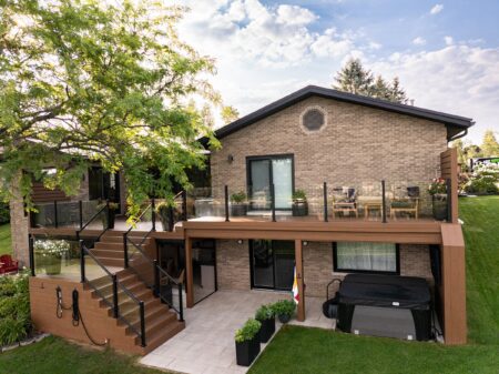 Two-story brick house with glass balcony, patio, stairs, outdoor hot tub, and landscaped yard on a sunny day.