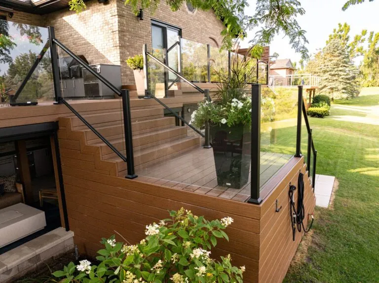 Modern raised wooden deck with glass railings, stairs, potted plants, and a view of a landscaped yard.