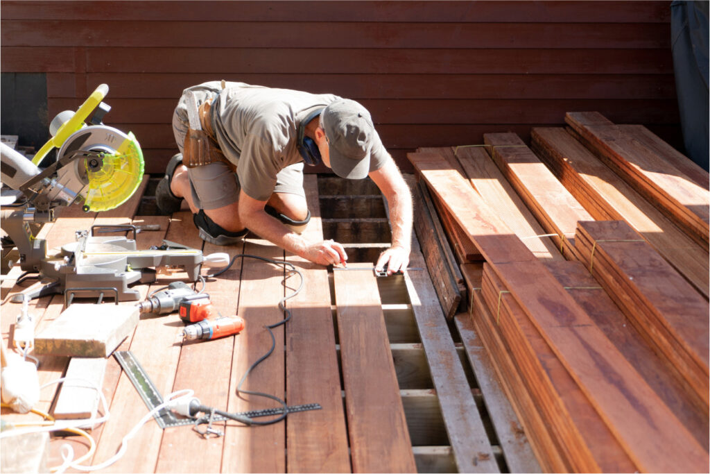 A person wearing a cap and tool belt is kneeling on a wooden deck under construction, measuring with a pencil and tape measure. Various tools, including a miter saw and drill, are nearby, and stacked planks are visible on the right.