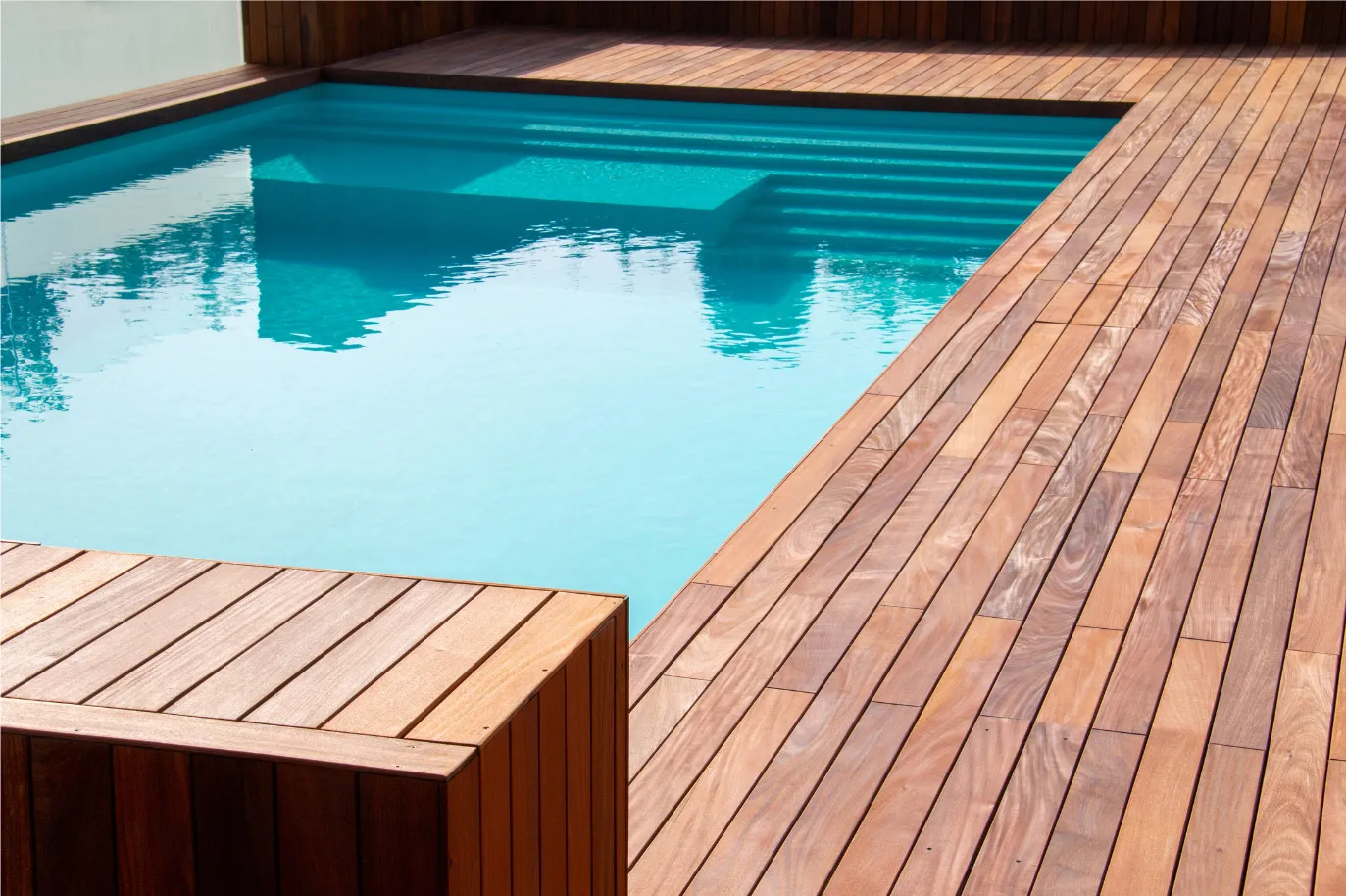 A modern swimming pool with clear blue water is bordered by a wooden deck featuring a bench. The pool steps are visible, and the surrounding area is sunlit, creating a serene and inviting outdoor space.